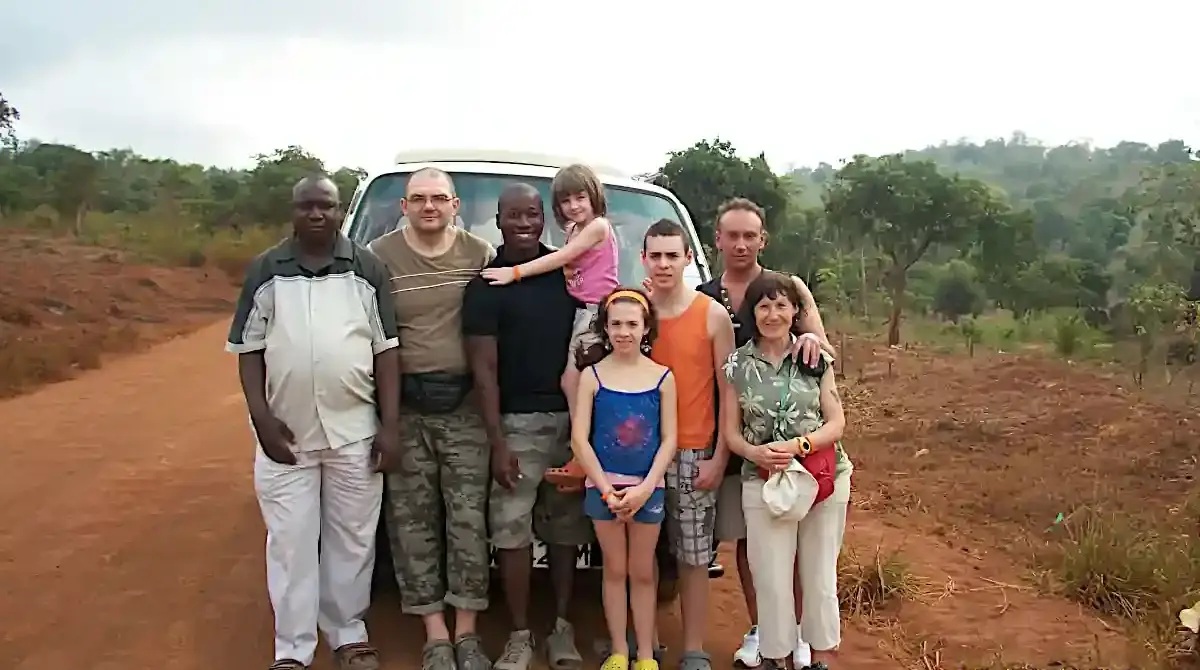 Safari familiare nello Tsavo con Wilderness 360: Morris tiene in braccio la figlia più piccola accanto al pulmino, con la famiglia sorridente nella terra rossa della savana.